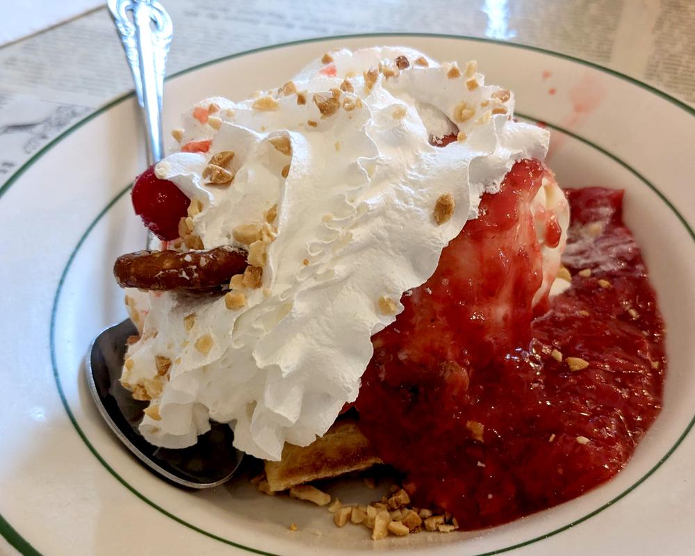 A waffle sundae with strawberry sauce, whipped cream, nuts, and a pretzel surrounding peanut butter ripple ice cream.